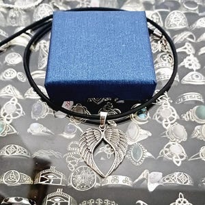 May include: A silver angel wing pendant necklace on a black cord. The necklace is displayed on a blue box and a background of silver rings.