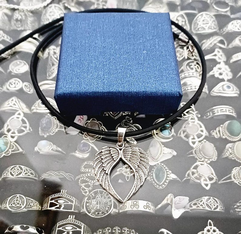 May include: A silver angel wing pendant necklace on a black cord. The necklace is displayed on a blue box and a background of silver rings.
