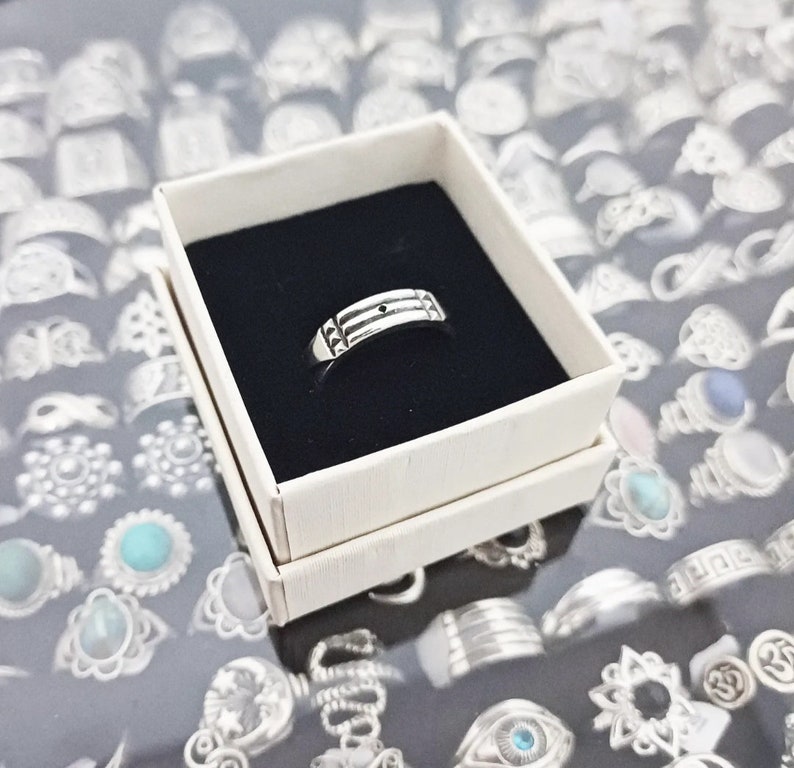 May include: A silver ring with a textured band and a small black dot in the center. The ring is displayed in a white box with a black interior.