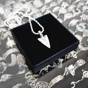 Puede incluir: Un collar de plata con un colgante de punta de flecha de plata en una caja de terciopelo negro. El collar se muestra en una mesa con otras joyas.