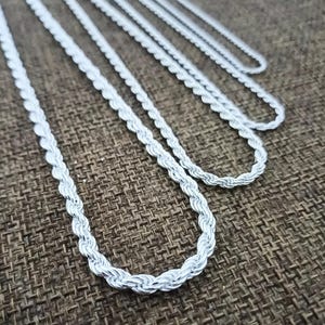 Può includere: Diverse catene a corda argentate di varie lunghezze sono esposte su uno sfondo marrone strutturato. Le catene sono disposte vicine, mostrando il loro design intrecciato e la superficie riflettente. L'immagine evidenzia la consistenza e la lucentezza dei gioielli.