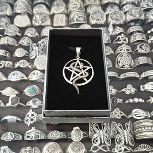 Pode incluir: Um pingente de metal prateado com uma estrela de cinco pontas dentro de um círculo, com uma cobra texturizada enrolada ao redor da estrela. O pingente está suspenso em um cordão preto e apresentado dentro de uma caixa preta retangular com forro de veludo.