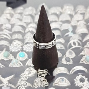 May include: A silver ring with a geometric design sits on a dark brown wooden ring display stand. The ring is surrounded by other silver rings.