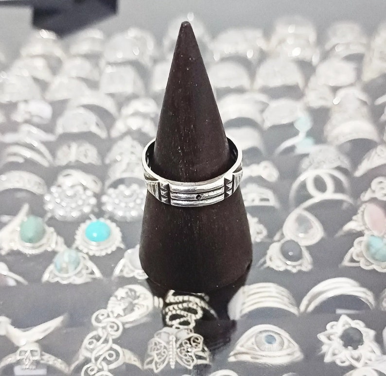 May include: A silver ring with a geometric design sits on a dark brown wooden ring display stand. The ring is surrounded by other silver rings.