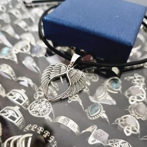 May include: A silver necklace with a heart-shaped pendant featuring two wings. The pendant is on a black cord and is displayed on a surface with other jewelry.
