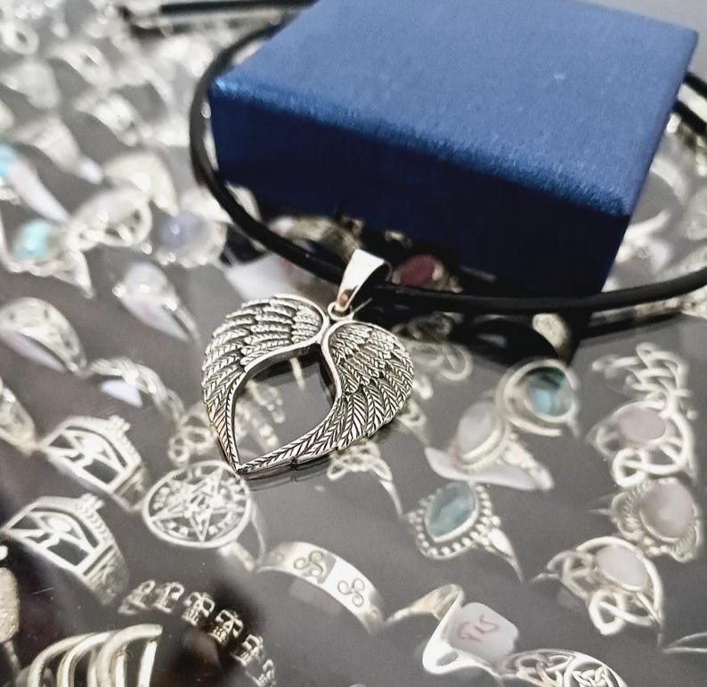 May include: A silver necklace with a heart-shaped pendant featuring two wings. The pendant is on a black cord and is displayed on a surface with other jewelry.