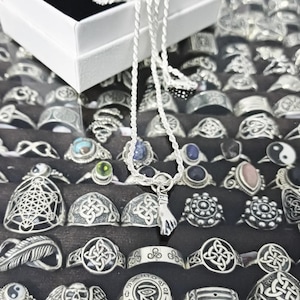 May include: A silver necklace with a hand pendant hangs in front of a display of various silver rings. The rings feature diverse designs, including Celtic knots, geometric patterns, and gemstone settings. A white box is visible in the upper left corner.