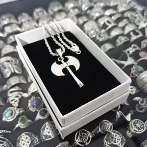 May include: A silver axe pendant necklace displayed in a white box with a black interior. The pendant is a detailed double-bladed axe design. The necklace is on a silver chain. The background shows a collection of silver rings.