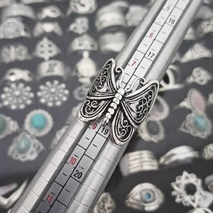 Puede incluir: Un anillo de plata con forma de mariposa con intrincados detalles de filigrana. El anillo se muestra en un medidor de anillos, con el tama&ntilde;o marcado como 9. El anillo est&aacute; rodeado de una variedad de otros anillos de plata.