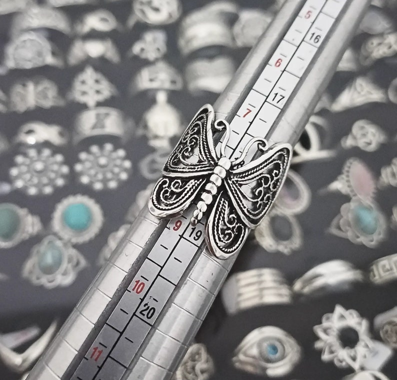 Puede incluir: Un anillo de plata con forma de mariposa con intrincados detalles de filigrana. El anillo se muestra en un medidor de anillos, con el tama&ntilde;o marcado como 9. El anillo est&aacute; rodeado de una variedad de otros anillos de plata.