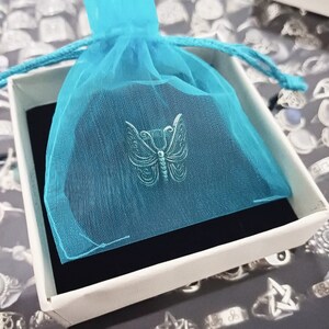 Puede incluir: Un dije de mariposa plateado en una bolsa de tul azul, sentado encima de una caja blanca con un forro de terciopelo negro.