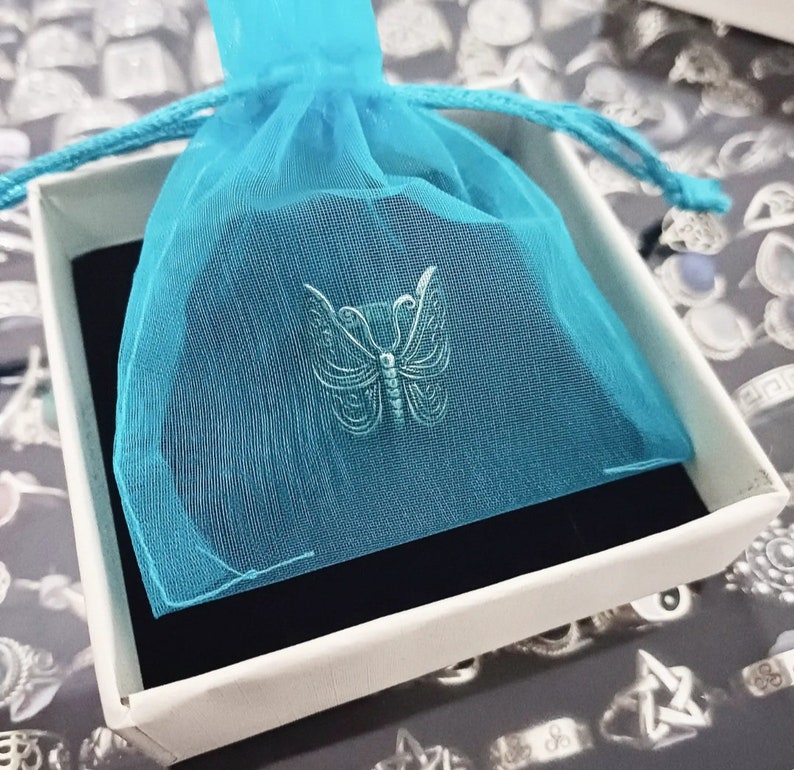Puede incluir: Un dije de mariposa plateado en una bolsa de tul azul, sentado encima de una caja blanca con un forro de terciopelo negro.