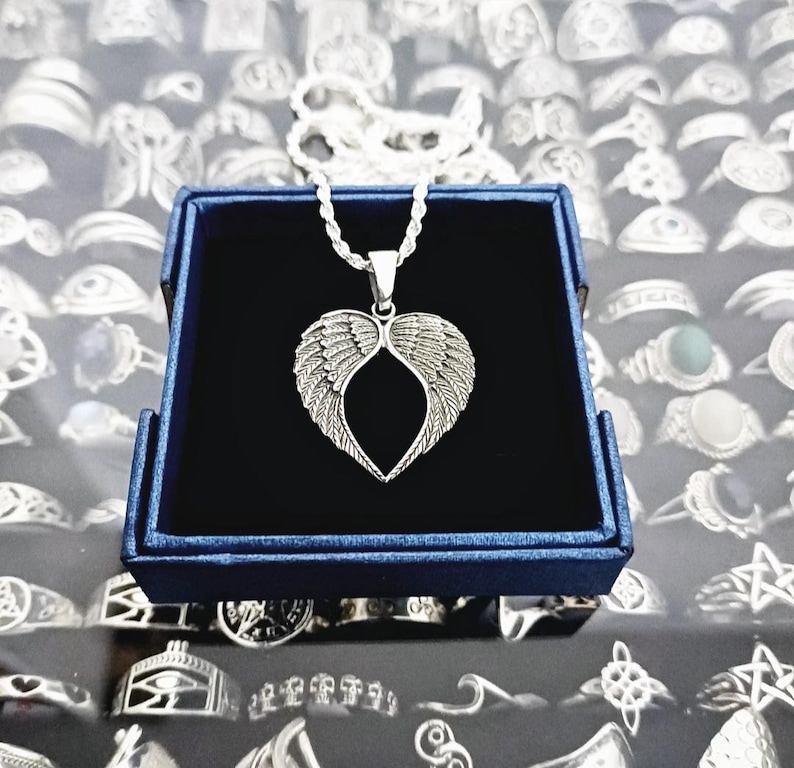 May include: A silver necklace with a heart-shaped pendant featuring angel wings. The necklace is displayed in a blue velvet jewelry box.