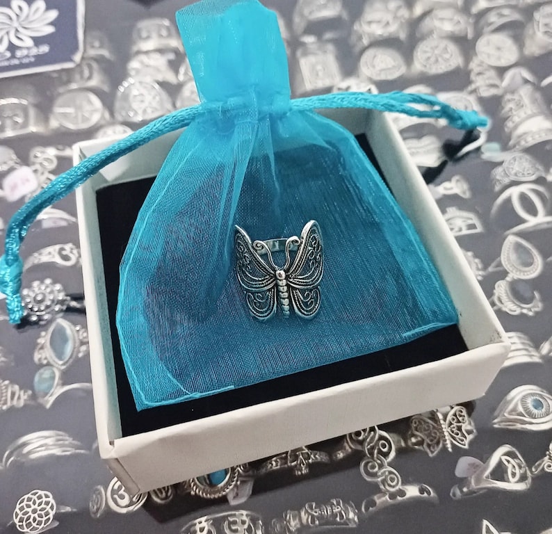 Puede incluir: Un anillo de mariposa plateado en una bolsa de organza azul turquesa. El anillo est&aacute; en una caja de regalo blanca. La caja est&aacute; sobre una superficie con muchos anillos plateados.