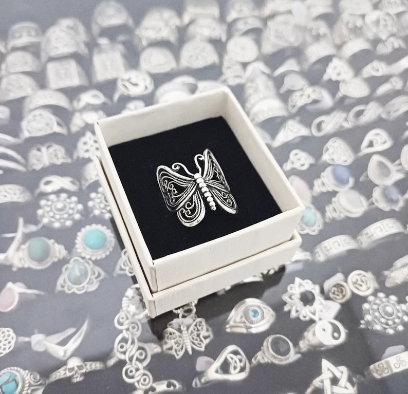 Puede incluir: Un anillo de plata con forma de mariposa en una caja blanca. El anillo est&aacute; rodeado de muchos otros anillos de plata.