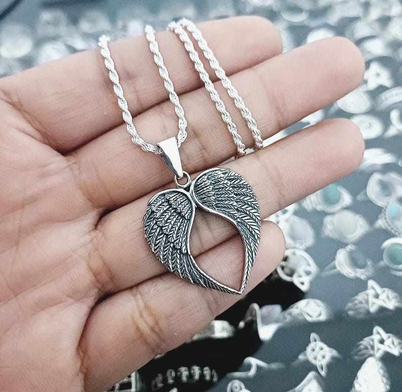 May include: A silver chain necklace with a pendant featuring a heart-shaped design with angel wings. The wings are detailed with intricate lines and have a textured finish.