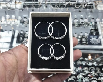 May include: Two pairs of silver hoop earrings with decorative details. The top pair has a beaded design, while the bottom pair has a more intricate design with small balls.