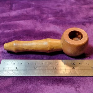 Puede incluir: Una pipa de madera con una cazoleta redonda y una boquilla larga y delgada. La pipa está hecha de madera marrón claro y mide aproximadamente 15 cm de largo.