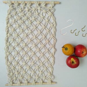 May include: A white macrame wall hanging with a wooden dowel at the top and bottom. The macrame is made with a knot pattern that resembles a diamond shape. There are five gold hooks on the right side of the image. Three red apples and one yellow apple are on the right side of the image.
