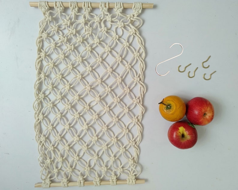 May include: A white macrame wall hanging with a wooden dowel at the top and bottom. The macrame is made with a knot pattern that resembles a diamond shape. There are five gold hooks on the right side of the image. Three red apples and one yellow apple are on the right side of the image.