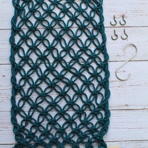 May include: A teal macrame wall hanging with a wooden dowel at the top and bottom. The macrame is made with a knot pattern that resembles a diamond shape. There are four silver S-shaped hooks to the right of the macrame.