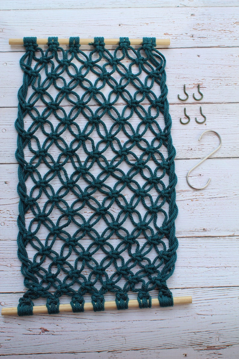 May include: A teal macrame wall hanging with a wooden dowel at the top and bottom. The macrame is made with a knot pattern that resembles a diamond shape. There are four silver S-shaped hooks to the right of the macrame.