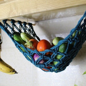 May include: A teal blue hanging hammock made of rope, filled with fresh produce, including cucumbers, tomatoes, and an eggplant. The hammock is hanging from a wooden cabinet.