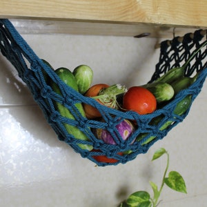 May include: A teal blue hanging produce hammock with cucumbers, tomatoes, and eggplant inside. The hammock is made of knotted rope and is hanging from a wooden shelf.