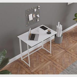 May include: A white desk with a laptop, tablet, and coffee cup. A black grid with photos and notes hangs on the gray wall. The desk has a minimalist design with white legs and a rectangular top. The floor is made of wood.