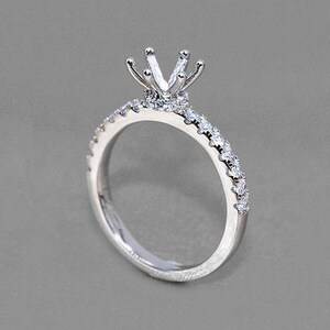 May include: A silver engagement ring with a prong setting. The band is set with small, sparkling stones. The ring's design features a crown-like setting to hold a centre stone, creating a classic and elegant look.