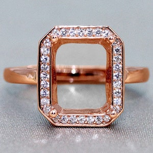 May include: A rose gold ring with a square-shaped setting surrounded by small clear stones. The setting is empty, ready for a gemstone.