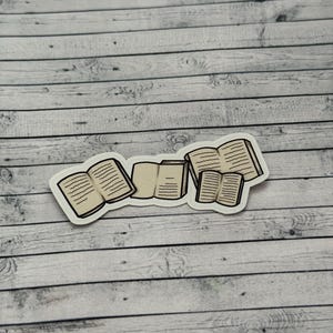 May include: A sticker depicting several open books with detailed pages. The books, in varying sizes and orientations, have a white border. The sticker is set against a light grey and white wood grain background.