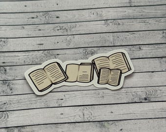 Op de afbeelding: Een sticker met een verzameling open boeken met gedetailleerde pagina's. De boeken zijn in verschillende maten en oriëntaties met een witte rand. De sticker is geplaatst tegen een lichtgrijze en witte houtnerf achtergrond.