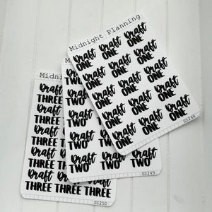 May include: A set of black and white stickers with the words "Draft One", "Draft Two", and "Draft Three" on them. The stickers are arranged in rows and columns on a white background.