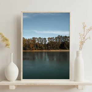 May include: A framed photograph of a river with trees lining the shore. The trees are in autumn colors, with shades of yellow, orange, and red. The water is calm and reflects the sky, which is a light blue with some clouds.