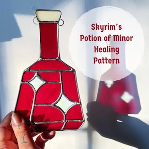 Può includere: Una finestra in vetro colorato appesa che rappresenta una bottiglia di pozione rossa con un tappo bianco e bordi argentati. La bottiglia ha un motivo geometrico con accenti bianchi. Il testo "Skyrim's Potion of Minor Healing Pattern" è scritto in bianco su un cerchio bianco.