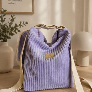 Könnte beinhalten: Eine hellviolette Umhängetasche aus Cord mit gerippter Textur, einem breiten, verstellbaren beigen Riemen mit Metallbeschlägen und einem kleinen, rechteckigen beigen Markenetikett, das mittig auf der Vorderseite angebracht ist.