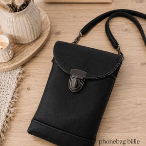 Könnte beinhalten: Eine schwarze, strukturierte Handytasche mit Klappverschluss, der durch einen dunklen Metalldrehverschluss gesichert ist, sowie einem langen Schulterriemen. Die Tasche hat eine rechteckige Form mit einer horizontalen Naht unten. Der Text lautet phonebag billie von Unikati.