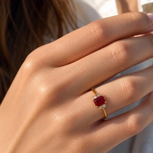 Puede incluir: Un anillo de oro con una piedra preciosa roja rectangular de corte esmeralda como piedra central, flanqueada por dos pequeñas piedras redondas transparentes a cada lado, sobre una fina banda de oro pulido.