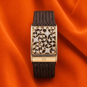 May include: A gold-toned bracelet with a dark brown woven strap. The rectangular centerpiece features a starburst design encrusted with small, sparkling gemstones. The letter "W" is engraved on the clasp.