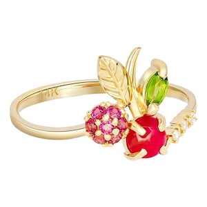 Puede incluir: Un anillo de oro con un diseño de cereza. El anillo presenta una cereza roja, una hoja verde y un grupo de piedras preciosas rosas. La banda es dorada con pequeñas piedras preciosas blancas. El anillo está marcado "14K".