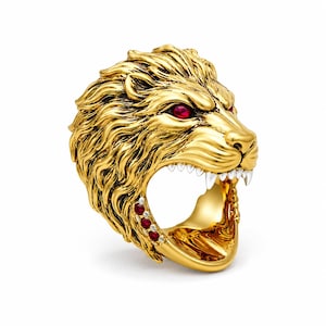Könnte beinhalten: Ein goldfarbener Ring in Form eines brüllenden Löwenkopfes mit detaillierter Mähne. Der Löwe hat zwei tiefrote Edelsteinaugen und drei kleinere rote Edelsteine an der Seite der Ringschiene. Im offenen Maul sind weiße Fangzähne zu sehen.