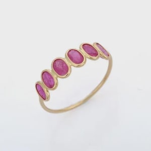 May include: A gold ring with six oval, bright pink gemstones set in a row. The gemstones are mounted in a gold band, creating a continuous design. The ring's polished gold band complements the bright pink stones.
