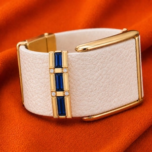 May include: A wide cuff bracelet featuring a textured off-white leather band with a polished gold-tone metal buckle. A vertical gold-tone bar is set with three rectangular blue gemstones and four small clear round stones as accents.
