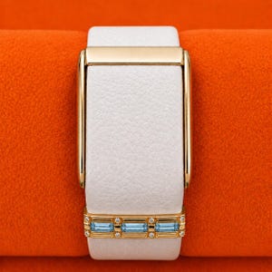 Puede incluir: Una correa de reloj de cuero blanco con un marco de metal dorado pulido y una barra decorativa dorada en la base, engastada con tres piedras preciosas rectangulares de color azul claro en talla baguette y pequeñas piedras transparentes.
