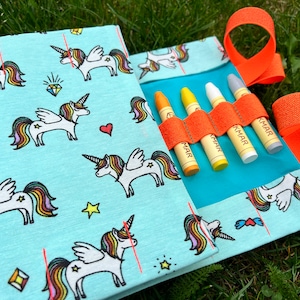 May include: A turquoise fabric pouch with a unicorn pattern and orange straps. The pouch has a blue flap that holds four crayons.
