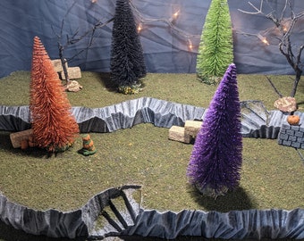 May include: A miniature Halloween scene with four small artificial Christmas trees in black, green, orange, and purple. The trees are arranged on a gray, rocky surface with a small set of stairs. There are also small, brown, square blocks scattered around the scene.