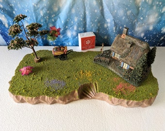 May include: A miniature village scene featuring a thatched-roof cottage, a tree, and a small cart. The scene is set on a green, grassy base with colorful flowers and a small staircase. A box with a snowflake logo is also present.