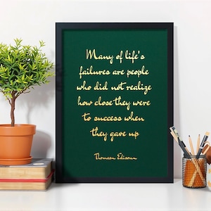 May include: A framed print with a green background and gold lettering displaying a quote by Thomas Edison. A small potted plant and books are on the left, with pencils and a small succulent on the right.