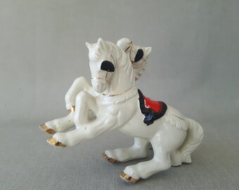 Puede incluir: Una figurita de porcelana blanca de un caballo de dos cabezas con una silla de montar roja y negra. El caballo está de pie sobre sus cuatro patas y tiene detalles dorados.